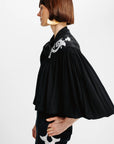 A model in profile wearing a black pleated blouse with white embroidered details on the shoulder and a matching black skirt with white embroidery, set against a plain white studio background.