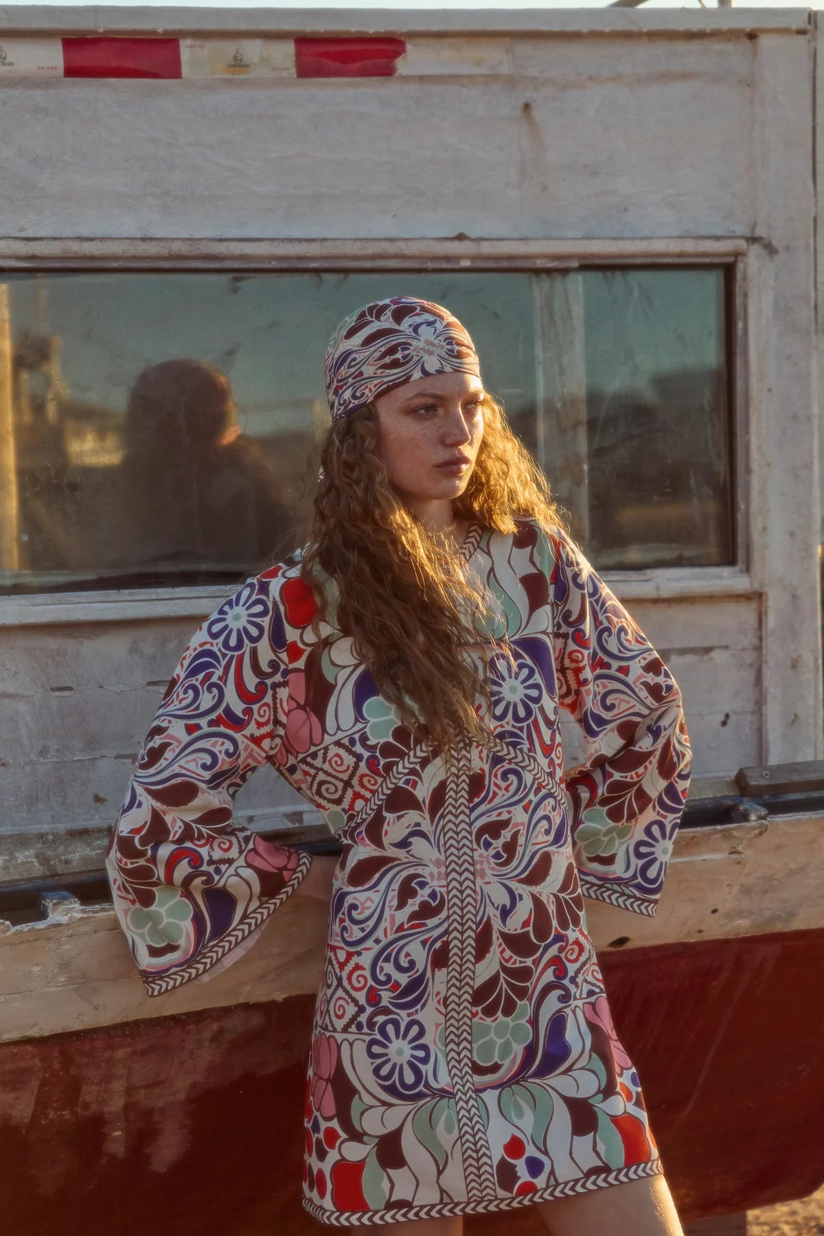 Woman wearing a colorful patterned dress and headscarf standing in front of a wooden structure.