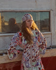 Woman wearing a colorful patterned dress and headscarf standing in front of a wooden structure.