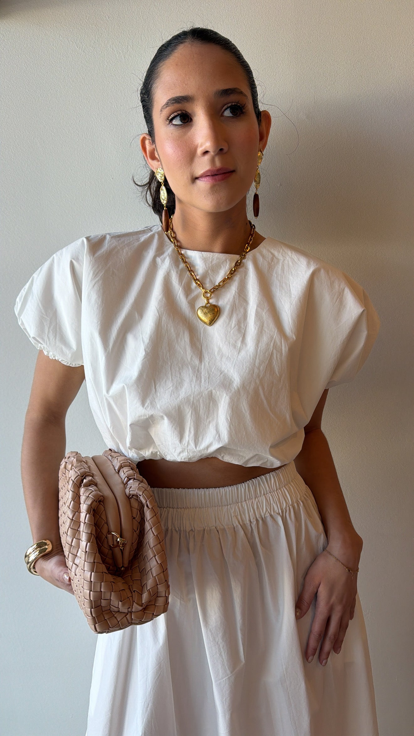 Woman wearing a white outfit with a gold necklace and holding a beige clutch against a plain background