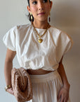 Woman wearing a white outfit with a gold necklace and holding a beige clutch against a plain background