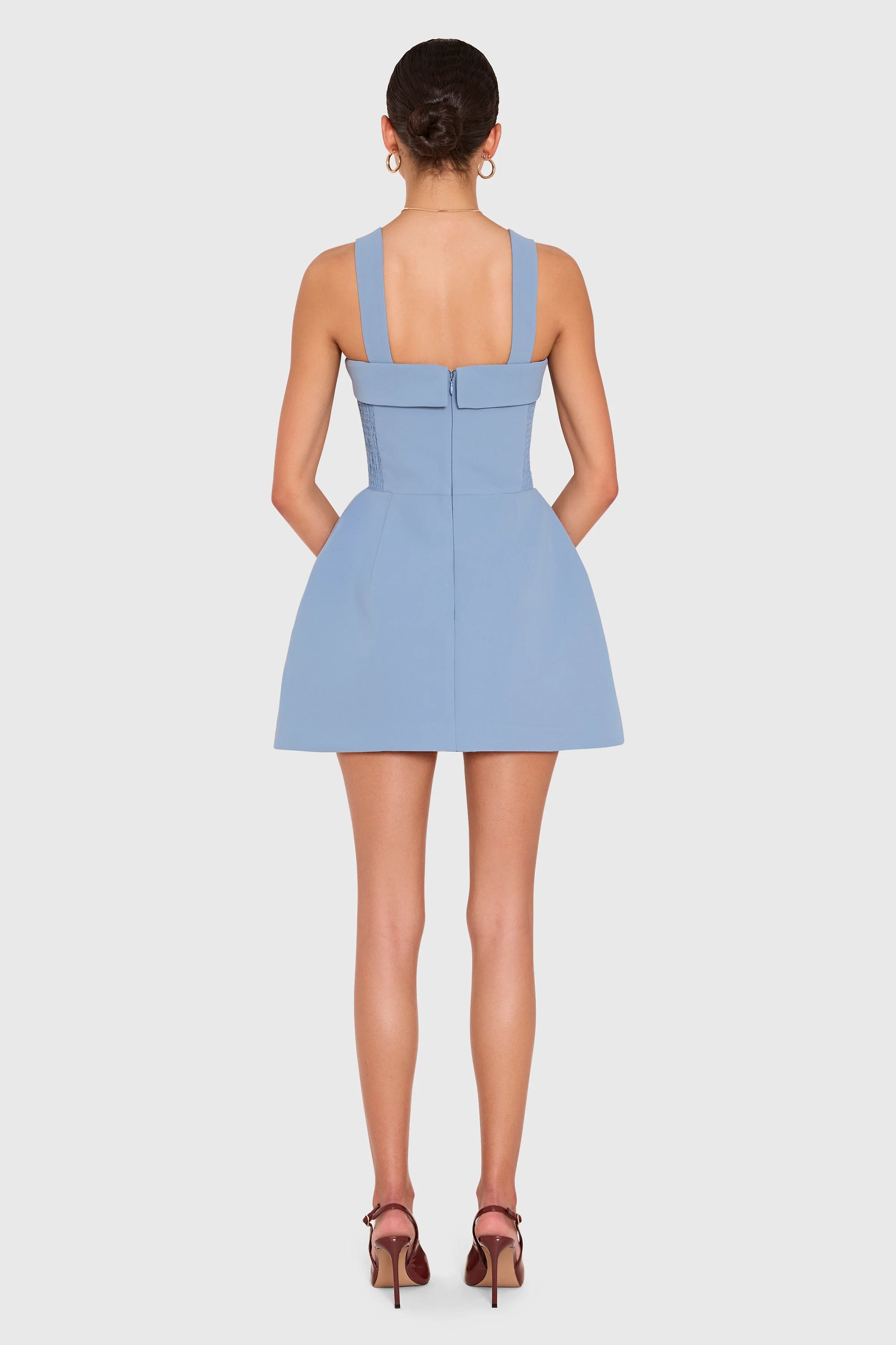 A model shown from the back wearing a light blue sleeveless mini dress with structured shoulder straps and a bow detail at the back, paired with brown high-heeled sandals against a light gray studio background.