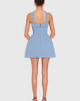 A model shown from the back wearing a light blue sleeveless mini dress with structured shoulder straps and a bow detail at the back, paired with brown high-heeled sandals against a light gray studio background.