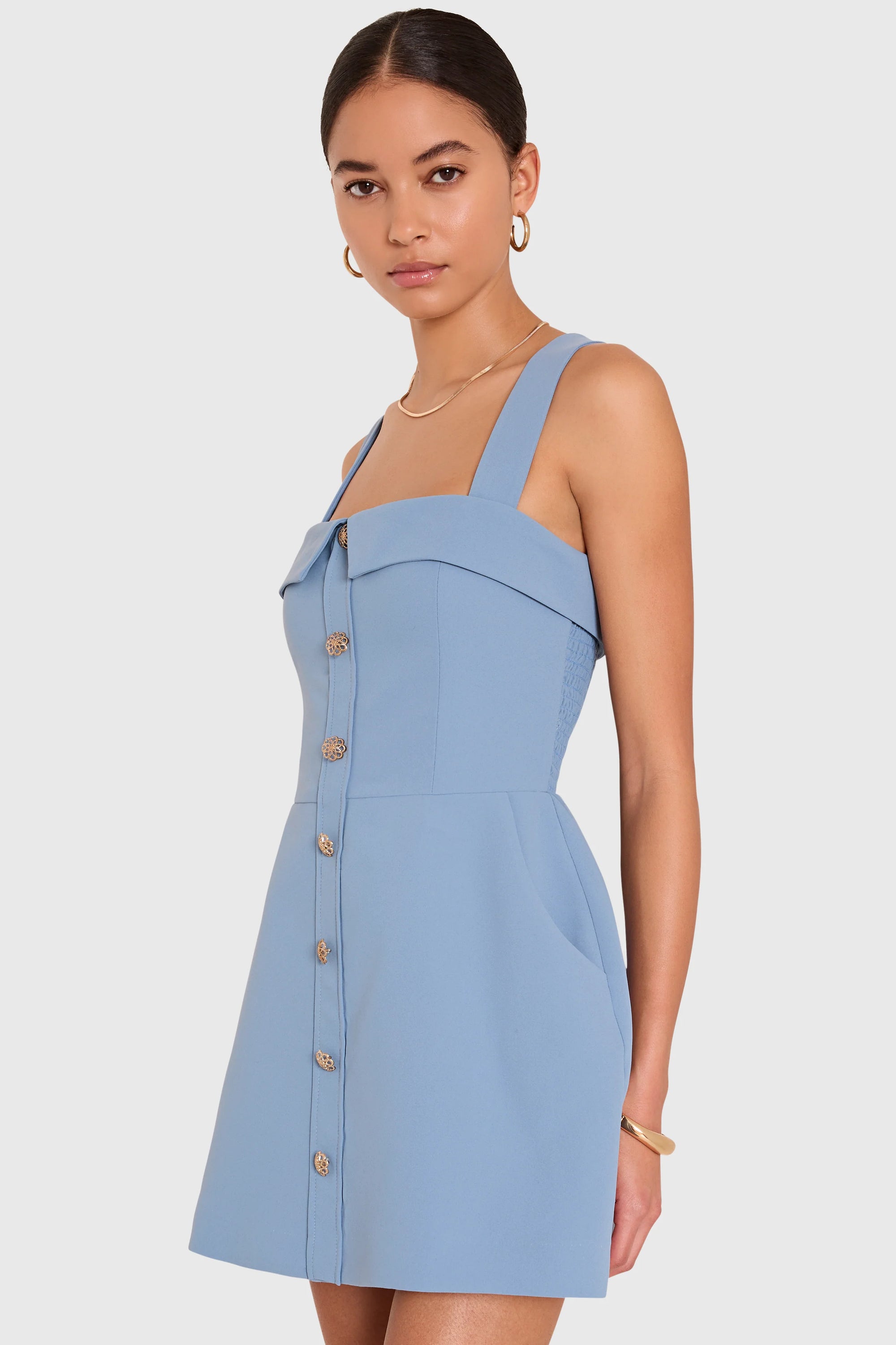 A woman modeling a light blue sleeveless mini dress with a structured, slightly draped bodice, square neckline, and a vertical row of gold buttons down the front, set against a plain light gray background.