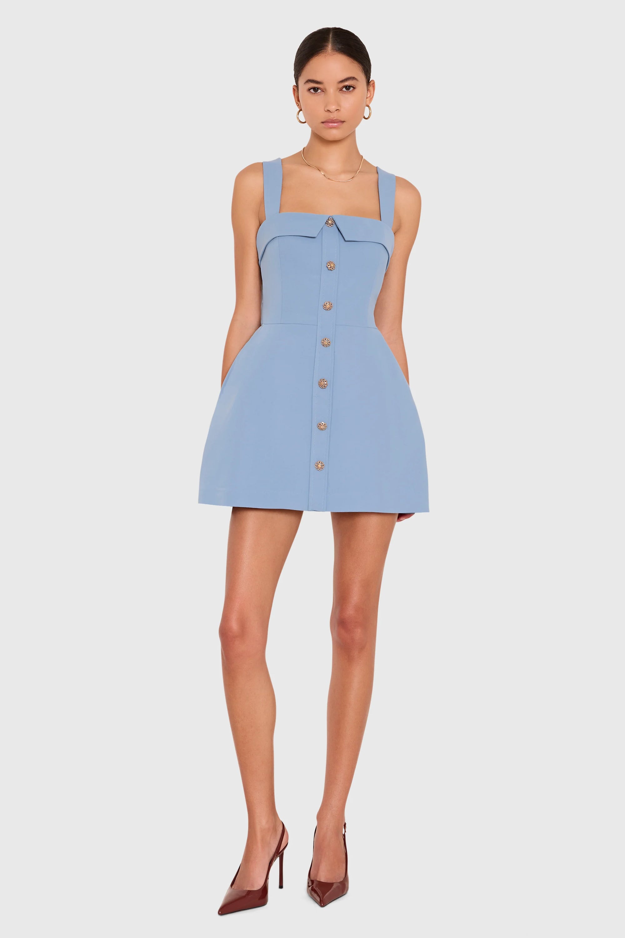 A model wearing a light blue sleeveless mini dress with a square neckline, button-front detail, and structured shoulders, paired with brown pointed-toe heels, against a light gray studio background.