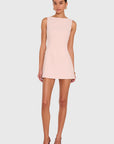 A woman modeling a sleeveless, light pink mini dress with a boat neckline, paired with brown pointed-toe heels, against a solid light gray background.
