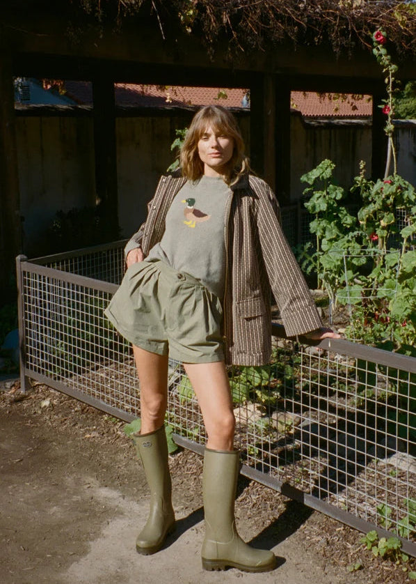 A woman stands outdoors in a garden setting wearing a light olive green graphic t-shirt, matching pleated shorts, a brown pinstriped cardigan, and olive green rubber rain boots.
