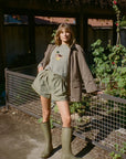 A woman stands outdoors in a garden setting wearing a light olive green graphic t-shirt, matching pleated shorts, a brown pinstriped cardigan, and olive green rubber rain boots.