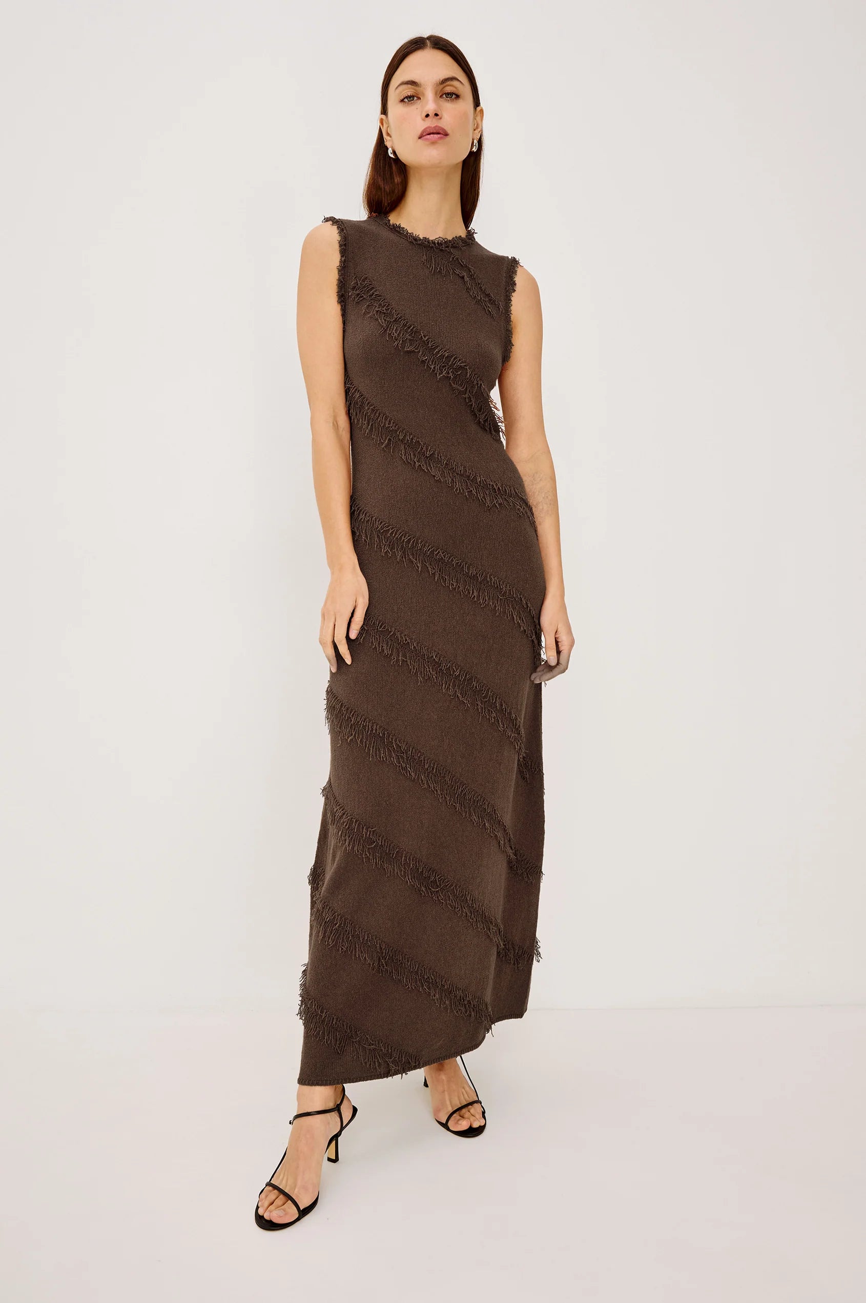 A woman modeling a sleeveless, high-neck, floor-length brown dress with diagonal textured fringe-like stripes, paired with black strappy heels, against a plain light gray studio background.