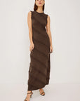 A woman modeling a sleeveless, high-neck, floor-length brown dress with diagonal textured fringe-like stripes, paired with black strappy heels, against a plain light gray studio background.