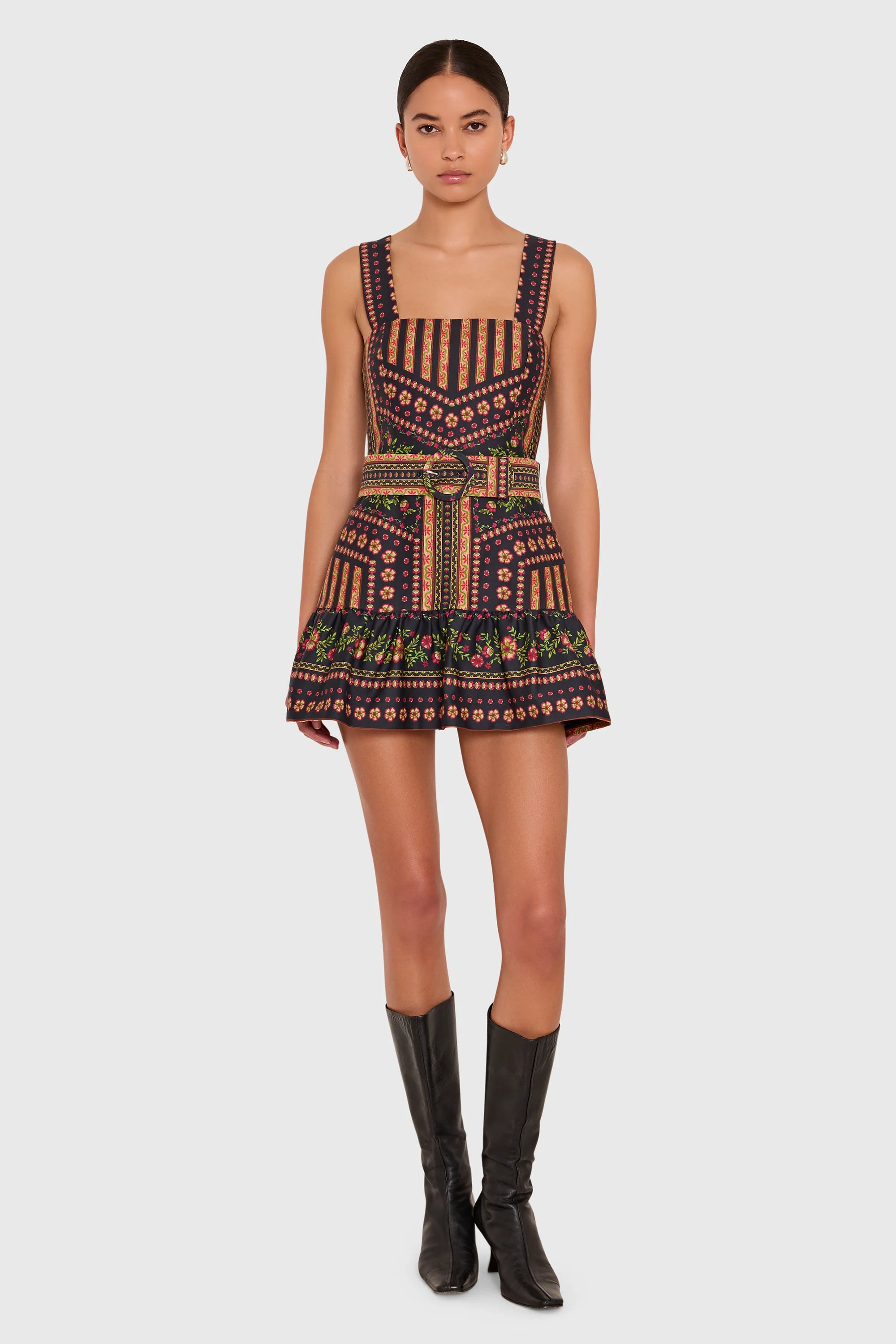 A model stands against a light gray studio background wearing a sleeveless mini dress with a geometric multicolor pattern and a matching tie belt, paired with black knee-high boots.
