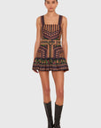A model stands against a light gray studio background wearing a sleeveless mini dress with a geometric multicolor pattern and a matching tie belt, paired with black knee-high boots.