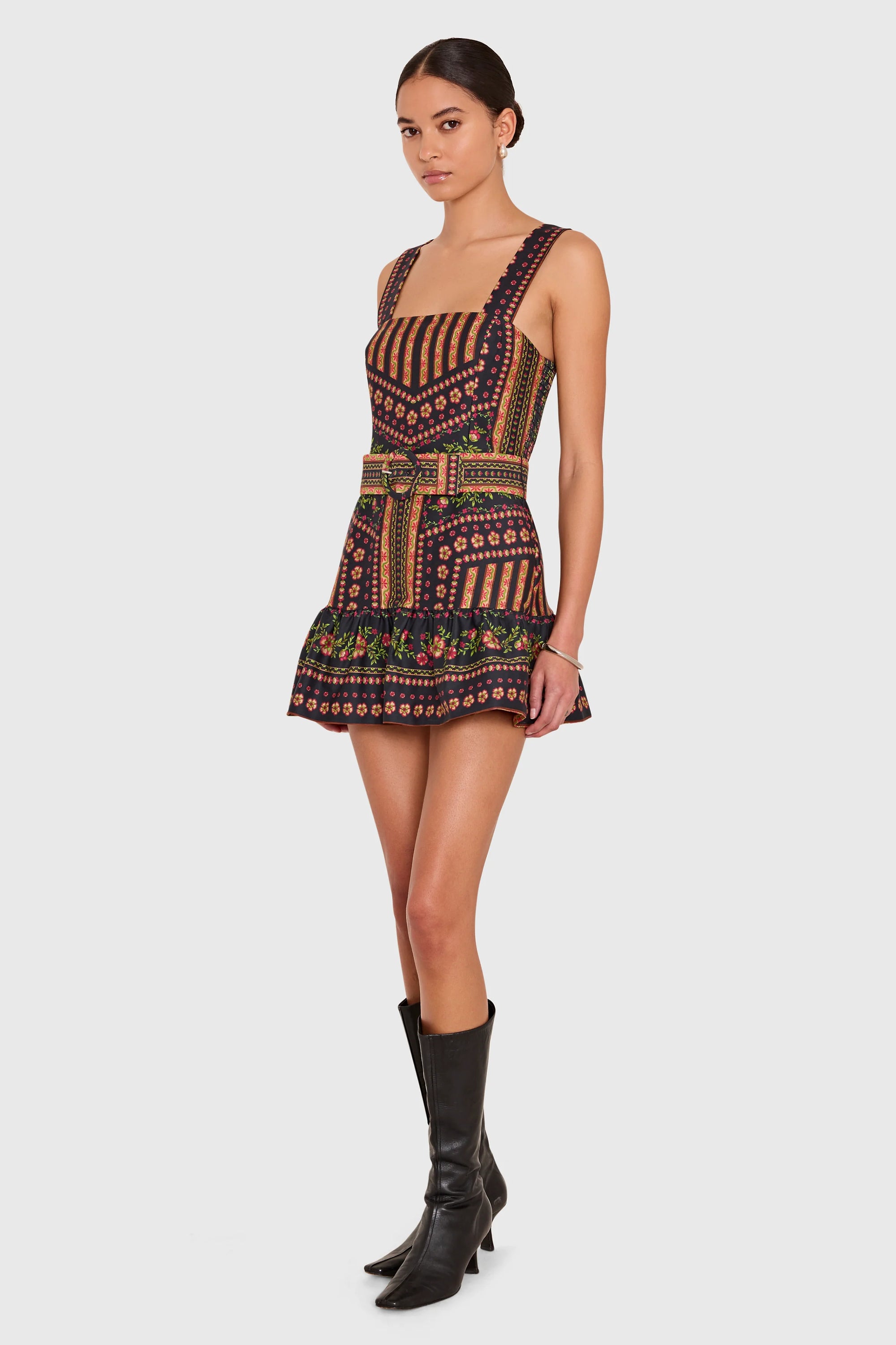 A model wearing a sleeveless mini dress with a geometric tribal pattern in brown, black, and orange, paired with black leather knee-high boots, set against a solid light gray background.