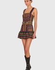 A model wearing a sleeveless mini dress with a geometric tribal pattern in brown, black, and orange, paired with black leather knee-high boots, set against a solid light gray background.