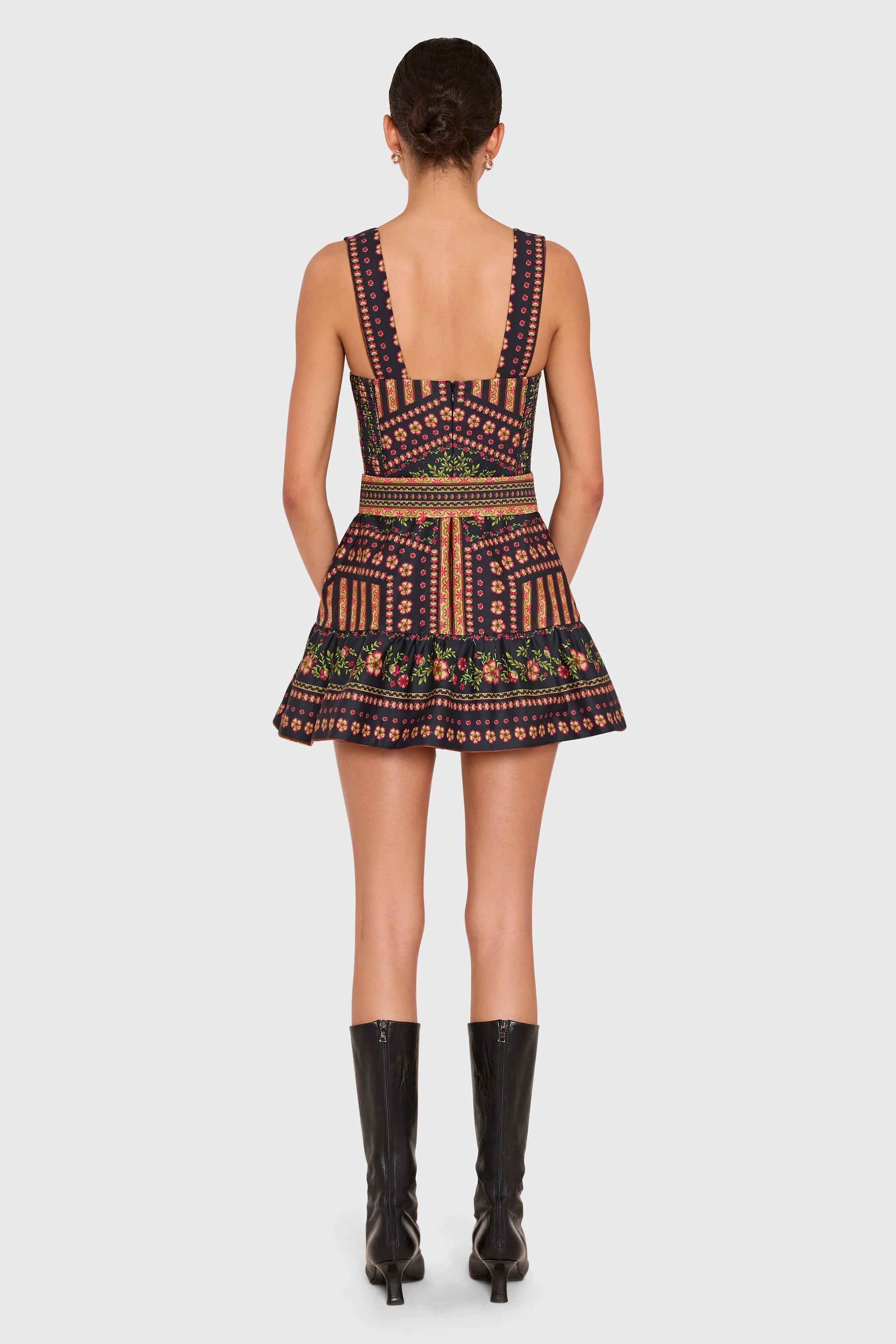 A model shown from the back wearing a sleeveless mini dress with a geometric pattern in navy, orange, and green, paired with black knee-high boots, against a light gray background.