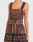 A sleeveless mini dress with a square neckline and wide shoulder straps, featuring a dark navy base with a colorful geometric and floral pattern in red, orange, green, and yellow. The dress includes a matching waist belt with a ring detail and is shown on a model against a light gray background.