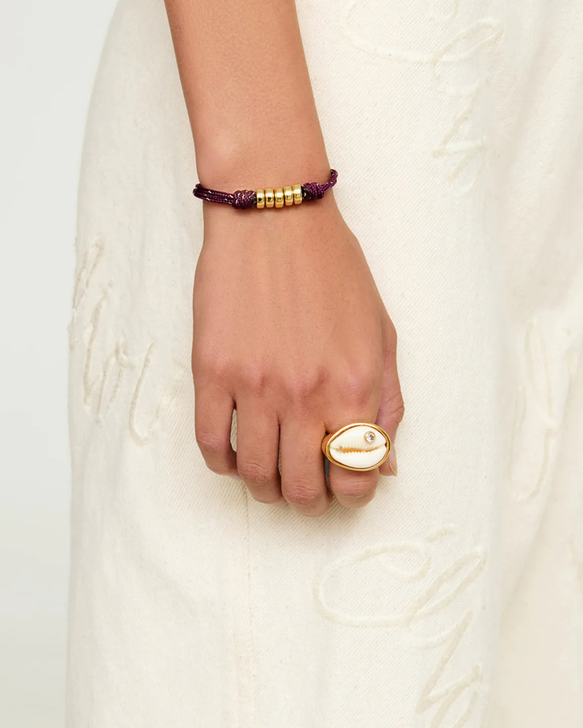 Hand wearing a gold ring and purple bracelet on a white background