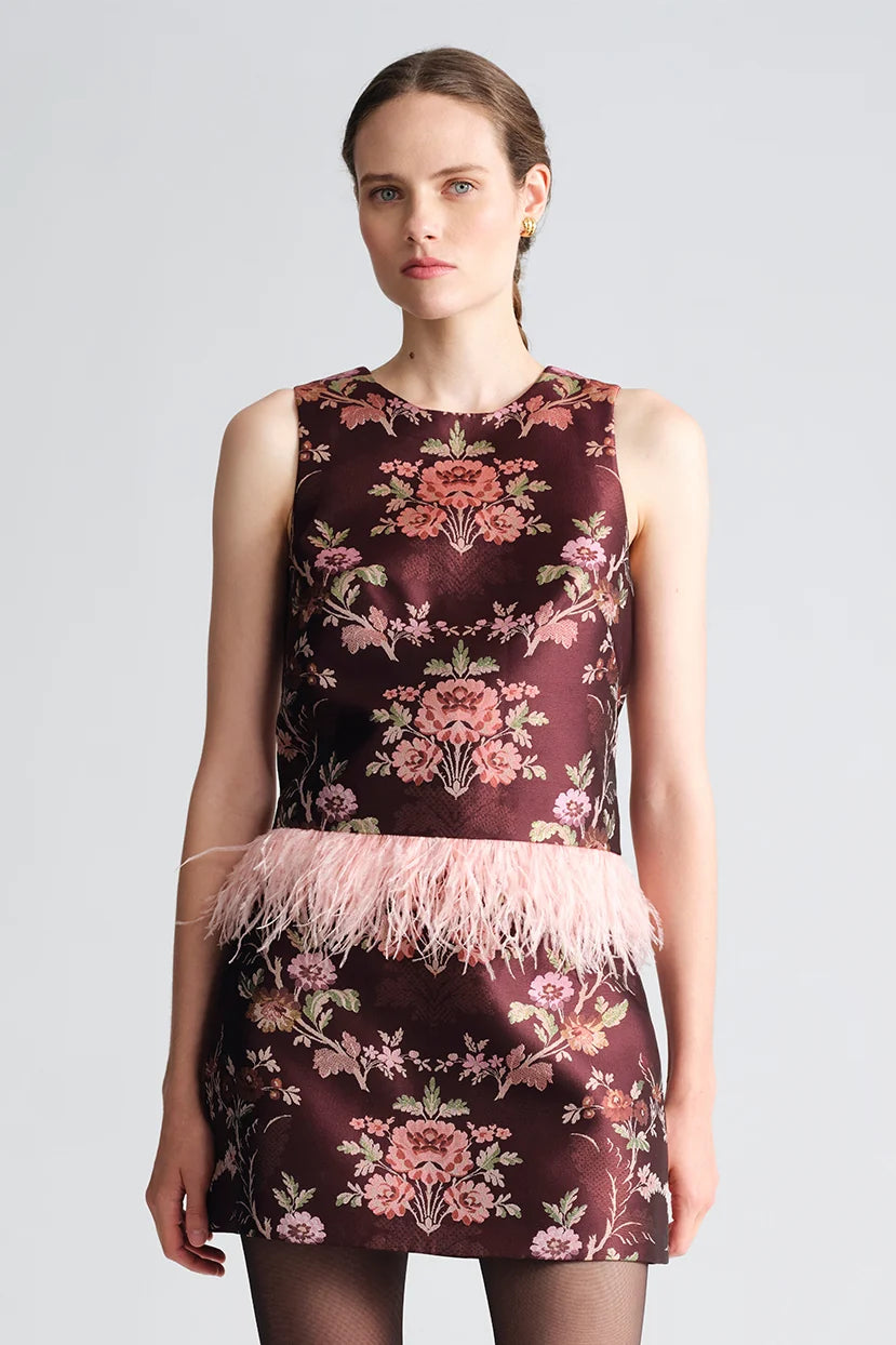 A sleeveless dark burgundy mini dress with a pink floral jacquard pattern and a light pink feather trim at the waist, modeled against a light gray background.