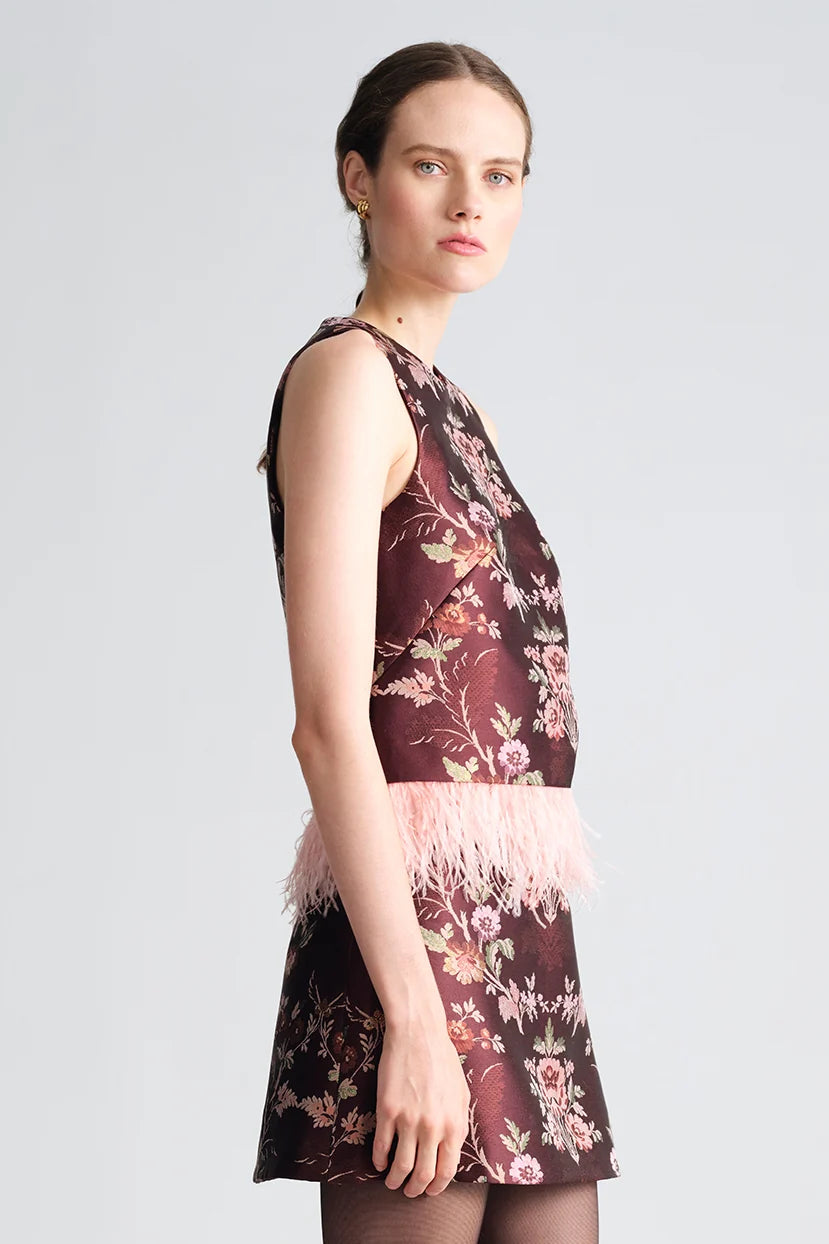 A woman modeling a sleeveless dark floral dress with a light pink feathered waistband, shown in profile against a light gray studio background.