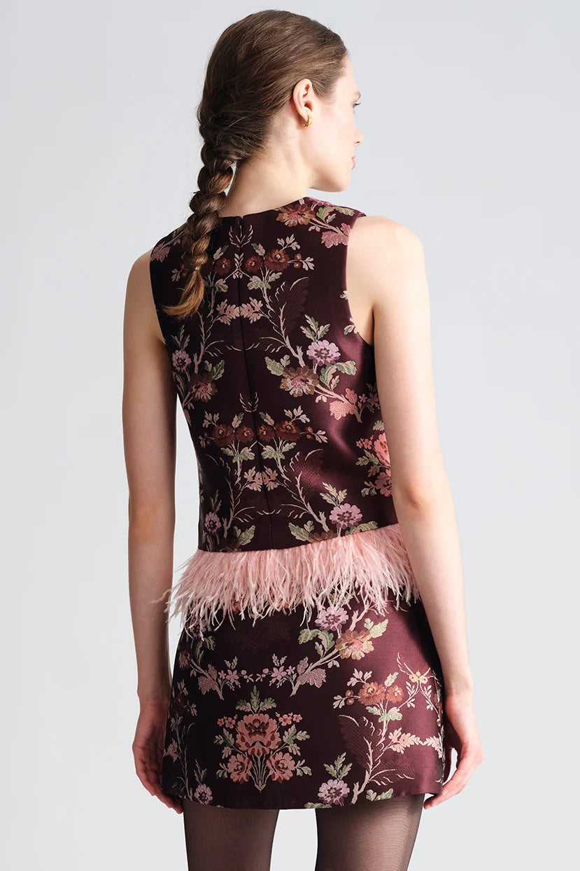 A woman shown from the back wearing a sleeveless dark burgundy mini dress with a pink floral brocade pattern and a light pink feather trim at the waist, set against a plain light gray studio background.