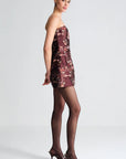 A model is shown in profile wearing a strapless mini dress with a floral pattern in burgundy and beige, paired with sheer black tights and dark brown pointed-toe heels, against a light gray studio background.