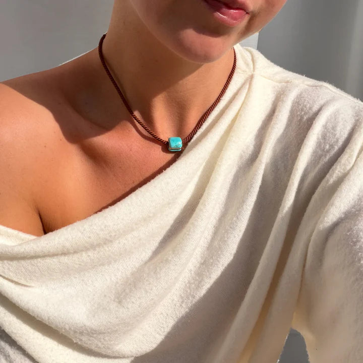 Person wearing a white garment and a necklace with a blue pendant against a neutral background