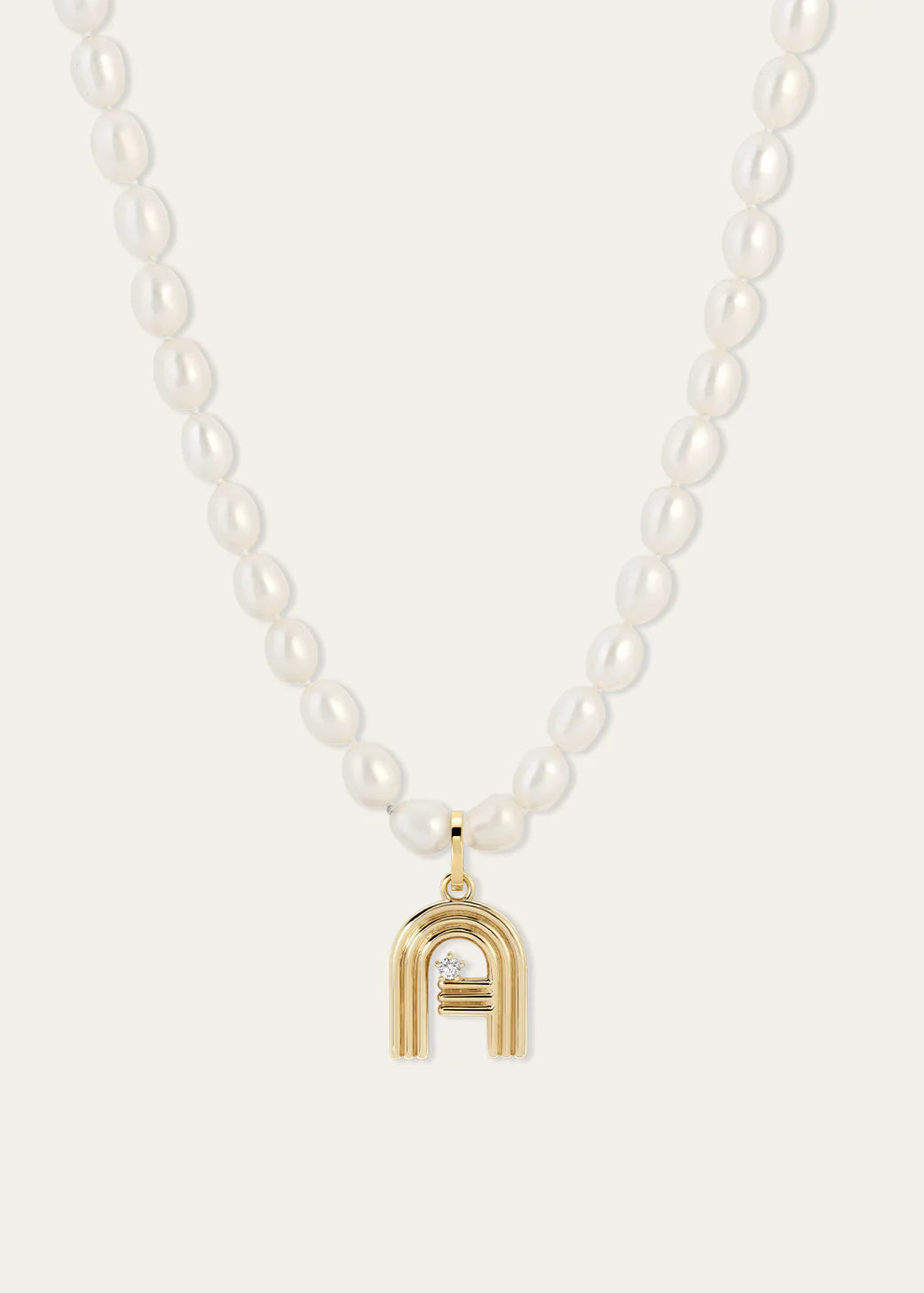 A white pearl beaded necklace with a gold pendant shaped like a stylized letter A, featuring a diamond in the center, set against a light beige background.