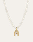 A white pearl beaded necklace with a gold pendant shaped like a stylized letter A, featuring a diamond in the center, set against a light beige background.