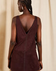 A woman shown from the back wearing a sleeveless deep burgundy mini dress with a plunging V-neckline and decorative stud detailing, set against a neutral beige curtain backdrop.