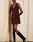 A model wearing a sleeveless burgundy corduroy mini dress with decorative studs down the front and black knee-high leather boots, posing in front of a beige curtain backdrop.