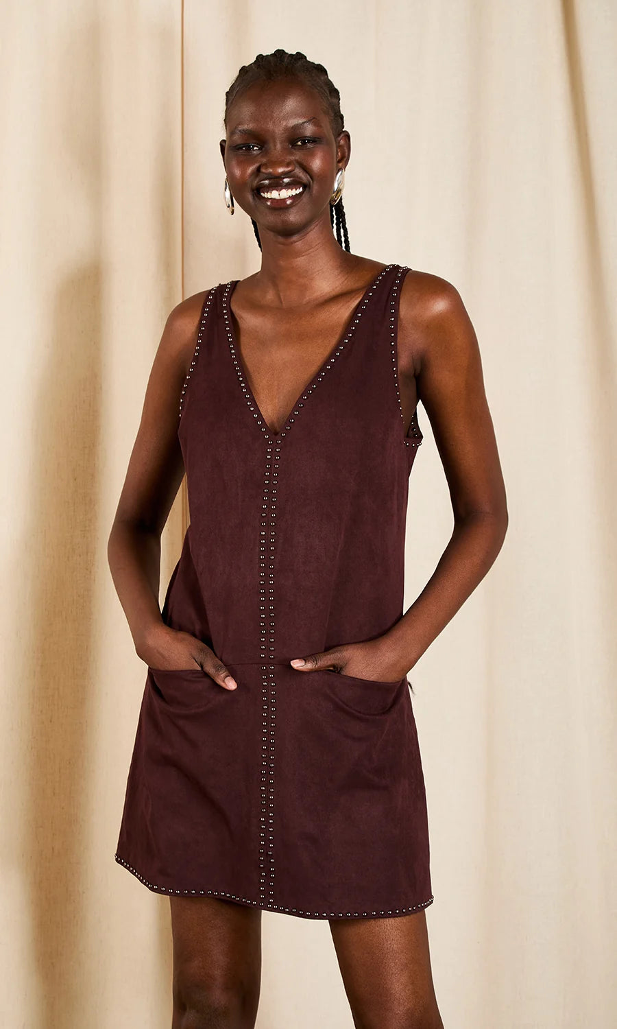 A woman modeling a sleeveless, deep burgundy dress with a V-neckline, decorative stud detailing along the neckline and front pockets, and a button-down front, set against a neutral beige curtain background.