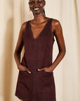 A woman modeling a sleeveless, deep burgundy dress with a V-neckline, decorative stud detailing along the neckline and front pockets, and a button-down front, set against a neutral beige curtain background.