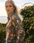 Woman wearing a colorful floral jacket outdoors with greenery and a beach in the background
