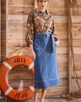 Woman in floral shirt and denim skirt standing next to an orange life buoy against a wooden background