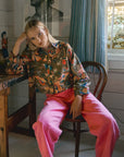 Person sitting on a chair wearing a colorful sweater and pink pants in a room with a painting and window.