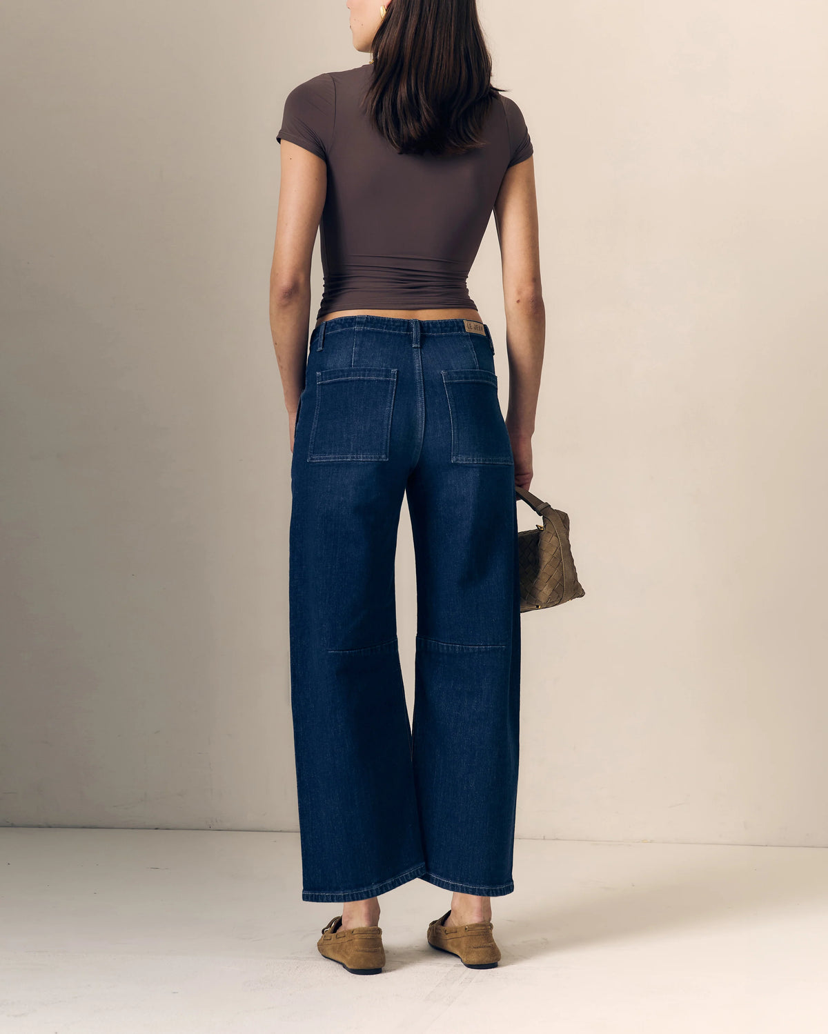 model waring blue jeans with a short sleeve brown top holding a woven bag on a cream background, back view 