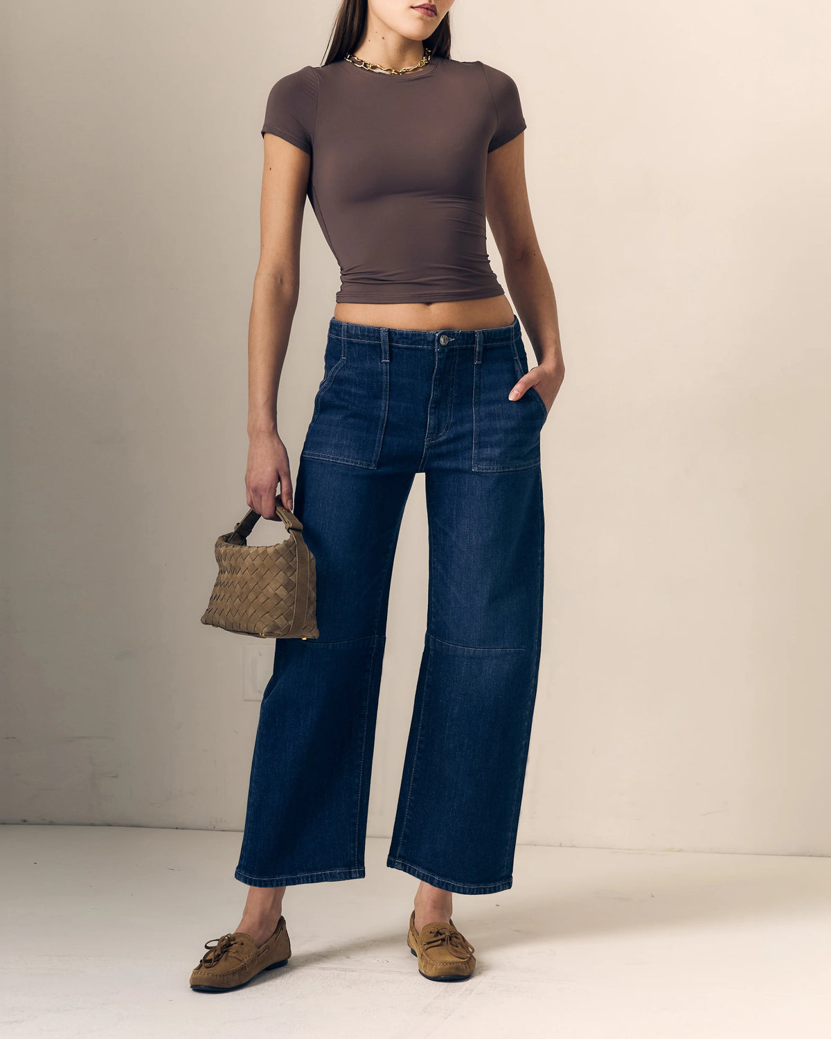 model waring blue jeans with a short sleeve brown top holding a woven bag on a cream background 