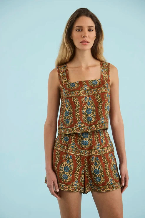A woman modeling a sleeveless two-piece romper set with a brown base and an intricate blue, green, and gold floral pattern, set against a solid light blue background.