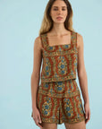A woman modeling a sleeveless two-piece romper set with a brown base and an intricate blue, green, and gold floral pattern, set against a solid light blue background.