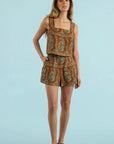 A woman modeling a sleeveless two-piece romper set with a brown, gold, and blue paisley pattern, paired with dark brown suede loafers, against a solid light blue background.