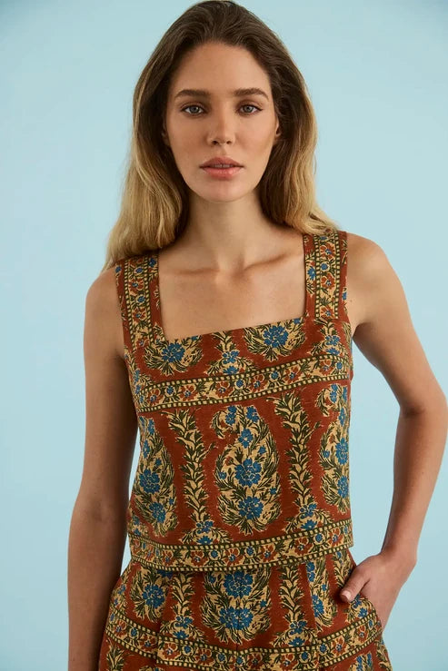 A woman modeling a sleeveless square-neck dress with an intricate bohemian-style floral and paisley print in rust red, blue, green, and gold, set against a solid light blue background.
