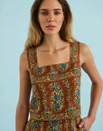 A woman modeling a sleeveless square-neck dress with an intricate bohemian-style floral and paisley print in rust red, blue, green, and gold, set against a solid light blue background.
