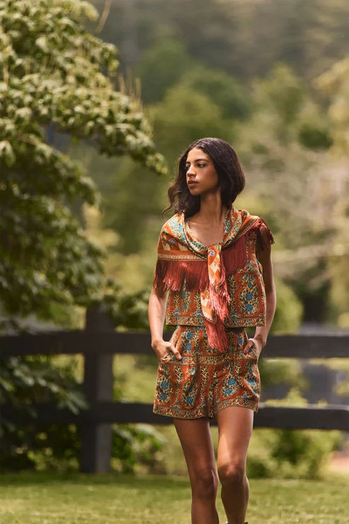 A woman wearing a bohemian-style two-piece matching set with a tie-front scarf, standing outdoors in a grassy park with trees and a fence in the background.