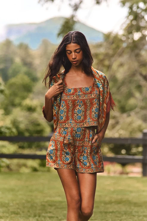 A woman wearing a bohemian-style two-piece outfit with a rust-orange, blue, and beige floral pattern, standing in a grassy outdoor setting with trees and a fence in the background.