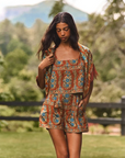 A woman wearing a bohemian-style two-piece outfit with a rust-orange, blue, and beige floral pattern, standing in a grassy outdoor setting with trees and a fence in the background.