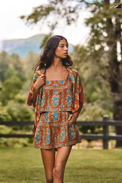 A woman stands outdoors in a grassy park wearing a bohemian-style two-piece outfit with a rust-orange base and blue floral patterns, featuring a matching short dress and a fringed shawl.