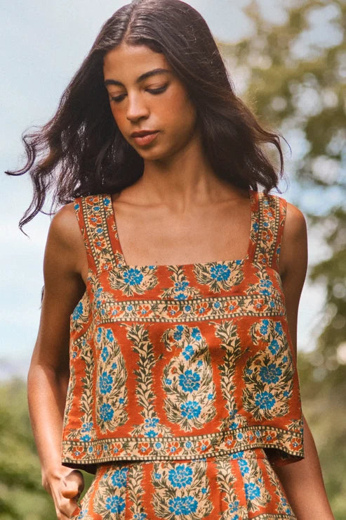 A woman wearing a sleeveless bohemian-style top and matching skirt set. The set features a rust-orange base color with intricate blue, green, and beige floral patterns.