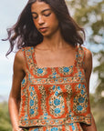 A woman wearing a sleeveless bohemian-style top and matching skirt set. The set features a rust-orange base color with intricate blue, green, and beige floral patterns.