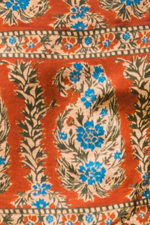 A close-up of a patterned textile featuring a rust-orange background with cream floral medallions, blue flower accents, and dark olive-green leafy motifs.