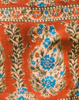 A close-up of a patterned textile featuring a rust-orange background with cream floral medallions, blue flower accents, and dark olive-green leafy motifs.
